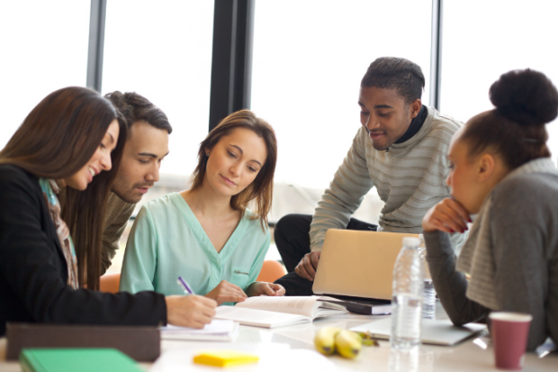 Photo of mulitple diverse people sitting around a conference table talking and looking at laptop