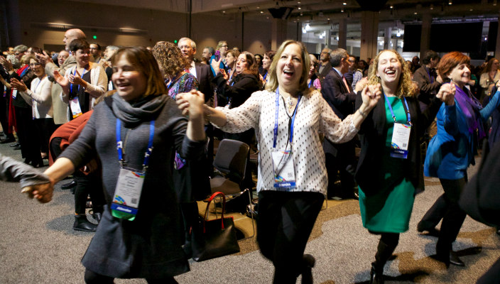 Women laughing as they hold hands and dance around the plenary room during a URJ Biennial song session