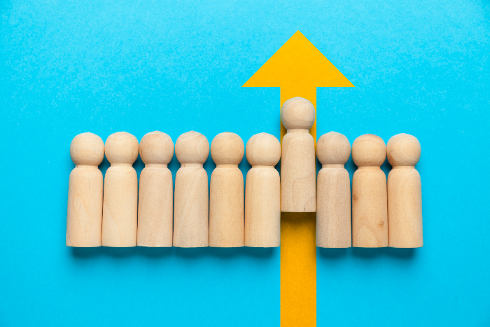 Line of wooden pegs shaped like people against a blue background with one peg moving upward against a yellow arrow as if to signify leadership