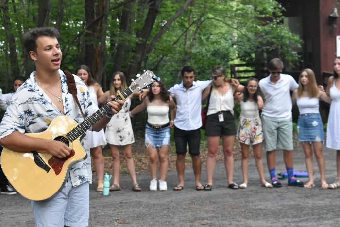 Summer camp staffer playing the guitar while standing in a circle of campers with their arms around one another