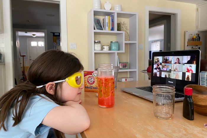 Young girl wearing googles while watching a Zoom screen surrounded by materials for conducting a science experiment 