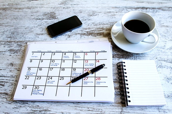 an image of a calendar on a desk, with a pen on top, and a cup of coffie and note pad next to it