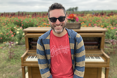 an image of Jay Rapoport, the director of URJ 6 Points Creative Arts Academy, standing in front of a piano outside in a garden