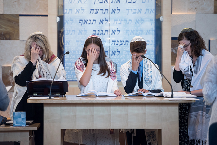 an image of four people standing on the bimah, two women, a boy and a girl, leading their b'nei mitzvah - all four of them are covering their faces with their hands as they pray. 