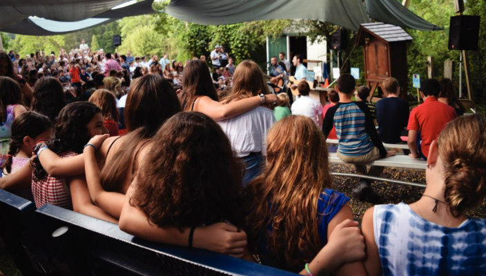Children with their arms around one another facing an outdoor bimah as at summer camp 