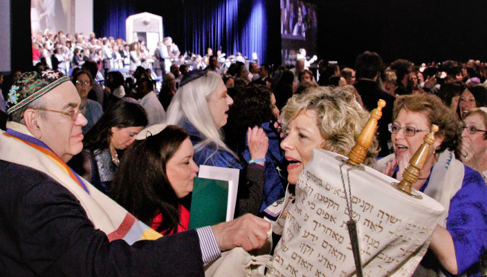 Smiling woman holding the Torah amongst a large crowd at the last URJ Biennial with the event stage visible in the background