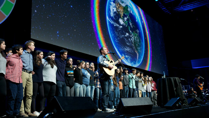 Jewish musician Dan Nichols plays guitar on stage with a line of teens behind him linking arms and a screen depicting the world above them