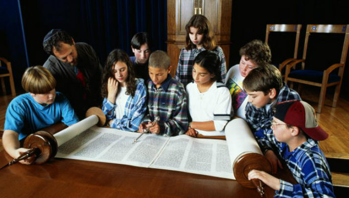 Young teens gathered around the bimah as they look at an open Torah scroll 
