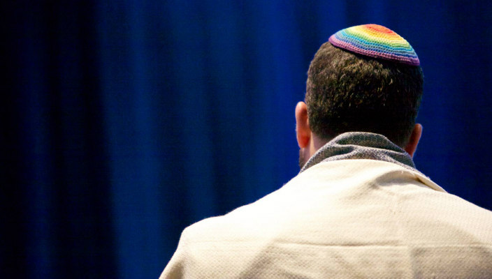 Back of a mans head wearing a rainbow kippah in front of a blue curtain background