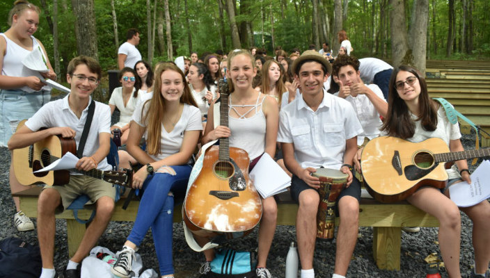 Group of songleaders sitting outdoors at a summer camp holding guitars and wearing all white as if on Shabbat