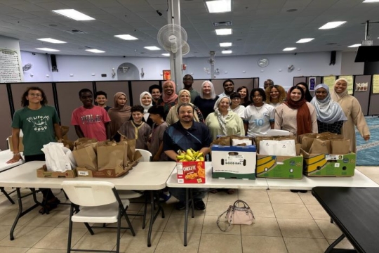 A smiling group of Muslim, Jewish, and Christian volunteers stand behind a table of food as they prepare to pack lunches for local children
